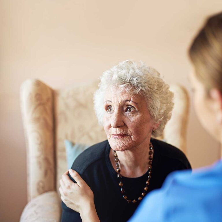 Adult child holding hands with an older parent while sitting together at a living room table.